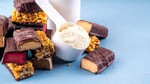 Assorted protein bars stacked next to a cup of protein powder on a gym surface