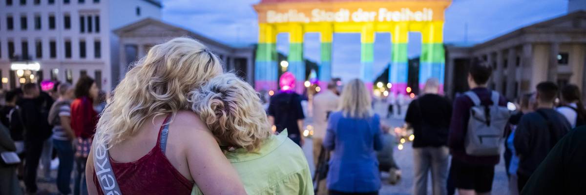 Mourners embrace near Brandenburg Gate illuminated in rainbow colors at Berlin Pride vigil