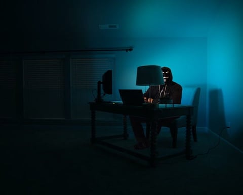An illustration of a masked hacker working at a computer in a dark room