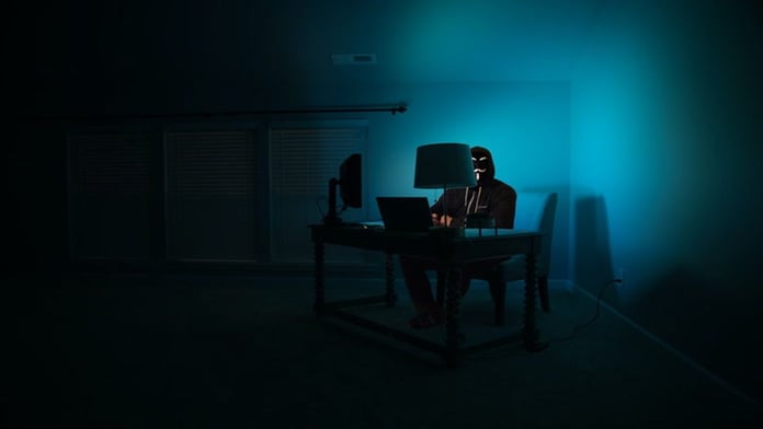 An illustration of a masked hacker working at a computer in a dark room