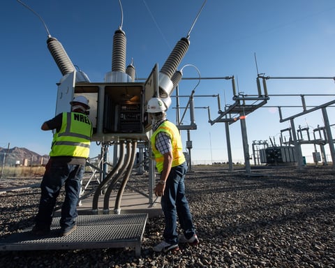 Technicians work at a high-voltage electrical substation with transformers and insulators