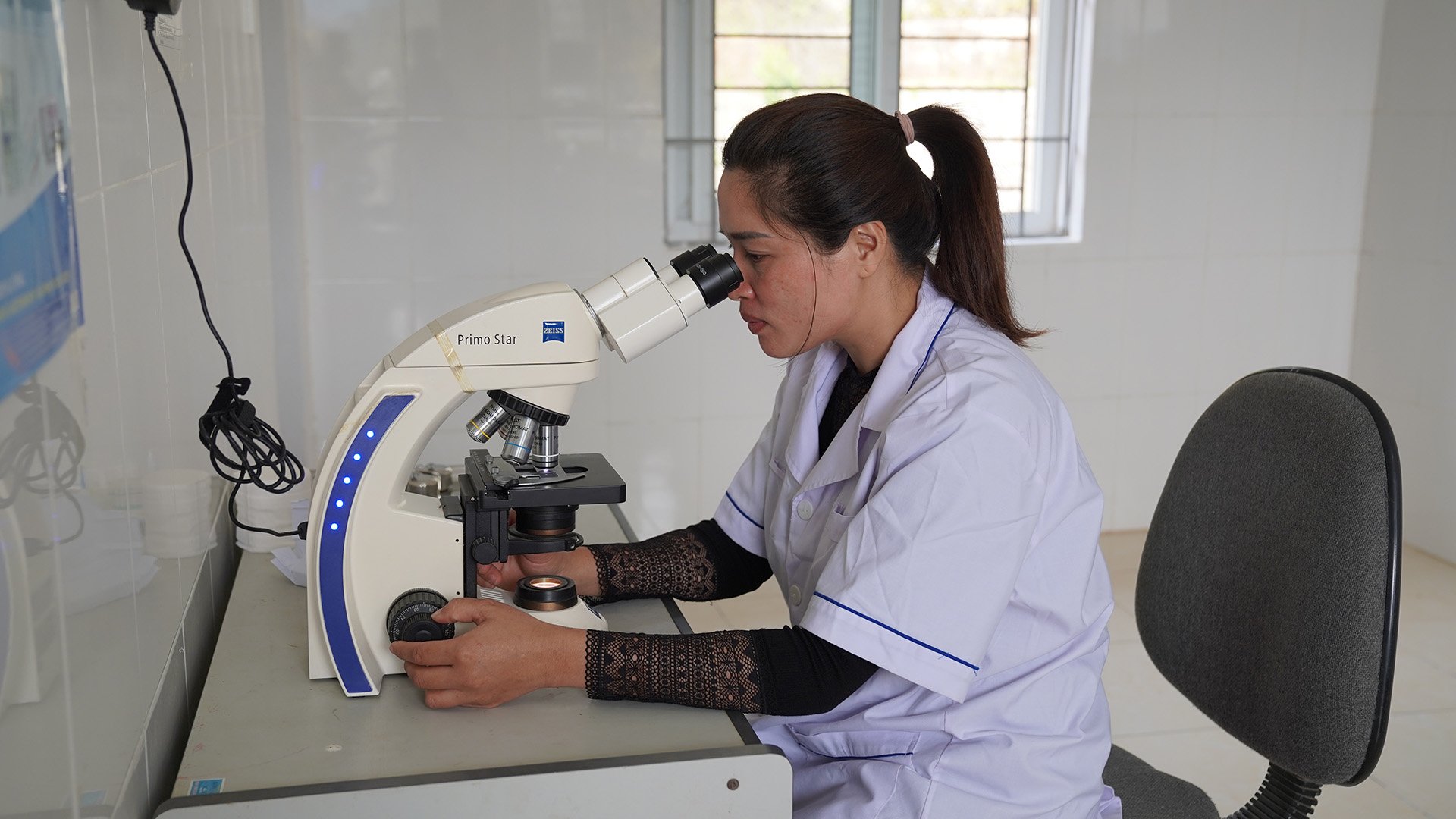A malaria laboratory officer examines a blood smear slide under a microscope to detect Plasmodium parasites in infected red blood cells