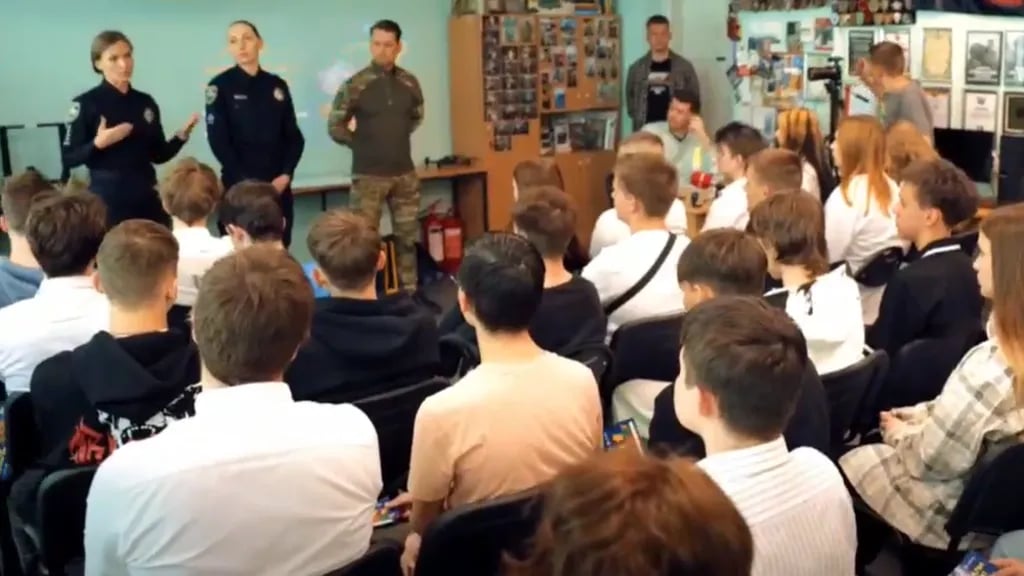 Law enforcement officers in uniform address a classroom of teenagers during a security awareness briefing on online recruitment and sabotage