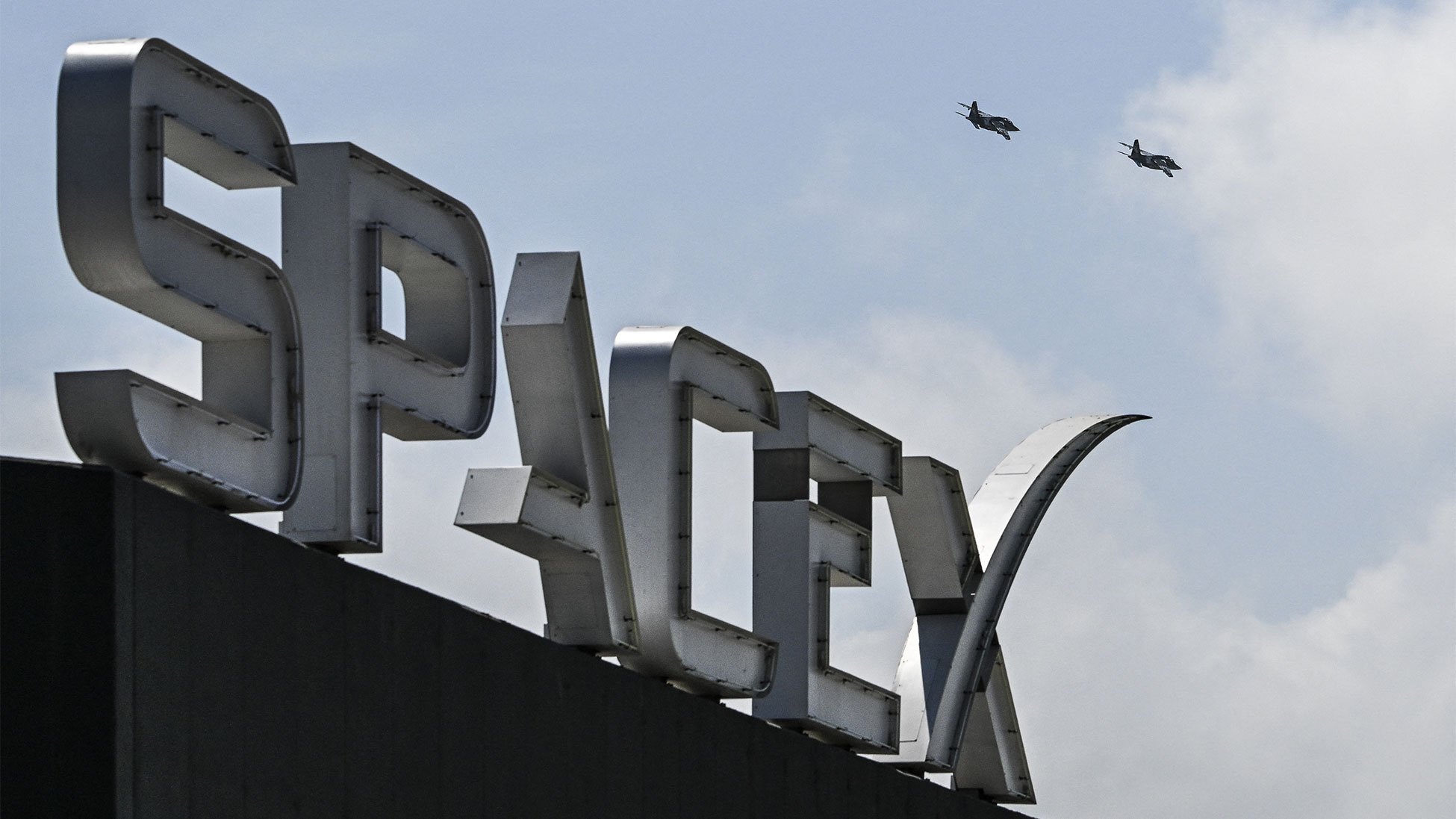 A fighter jet flies in formation near the SpaceX sign and Starship spacecraft at Starbase, Texas