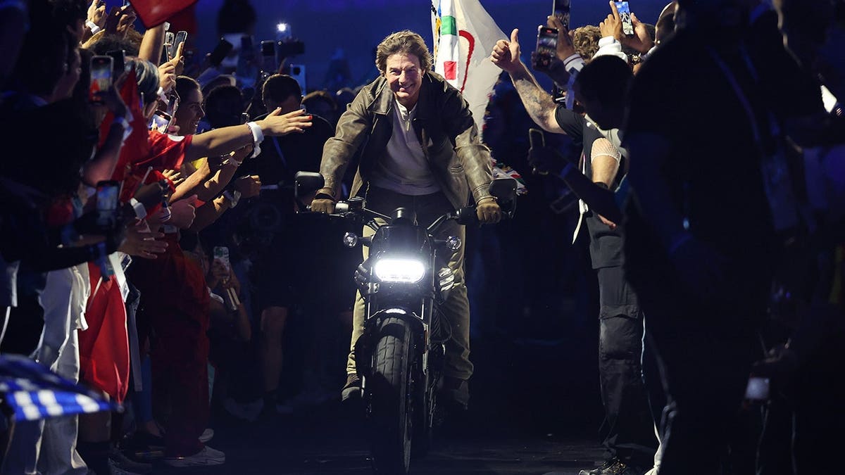 Tom Cruise on a motorcycle during a public appearance, with fans reaching toward him from the sidelines