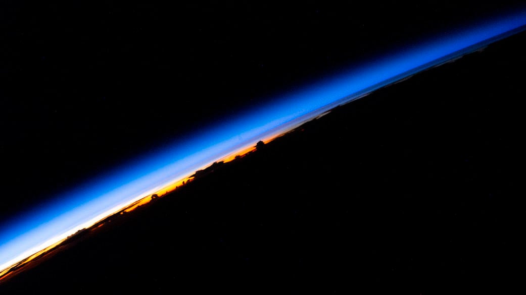 An orbital sunrise beams across Earth's horizon with silhouetted clouds below, seen from the International Space Station