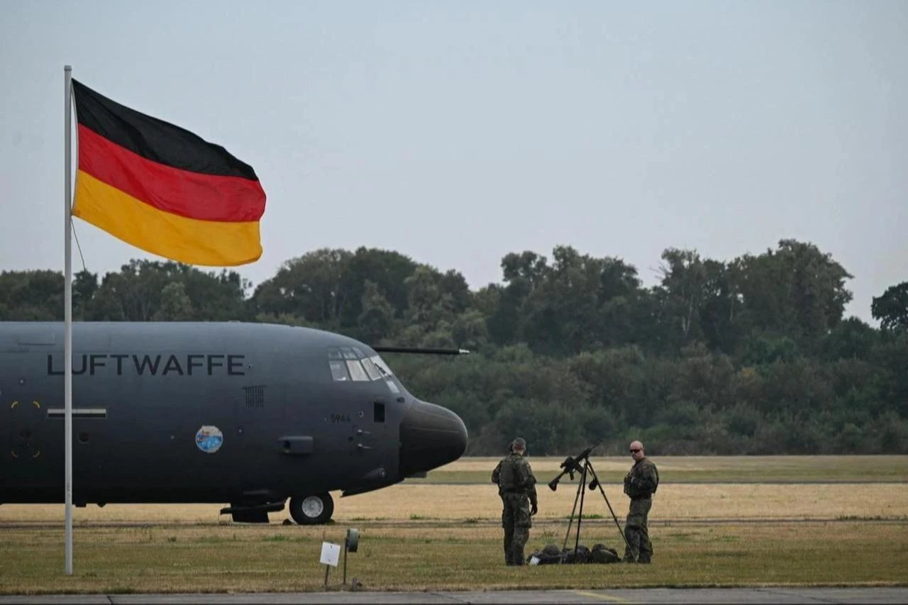 Two Bundeswehr soldiers stand beside a Luftwaffe C-130 transport aircraft on an airfield with the German flag flying