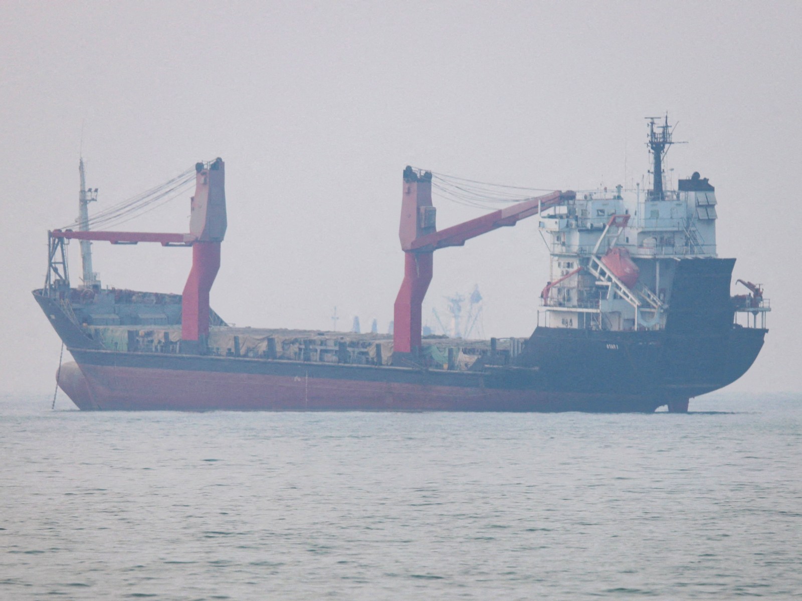 A general cargo ship with red deck cranes lies at anchor in flat grey water and haze near the Strait of Hormuz