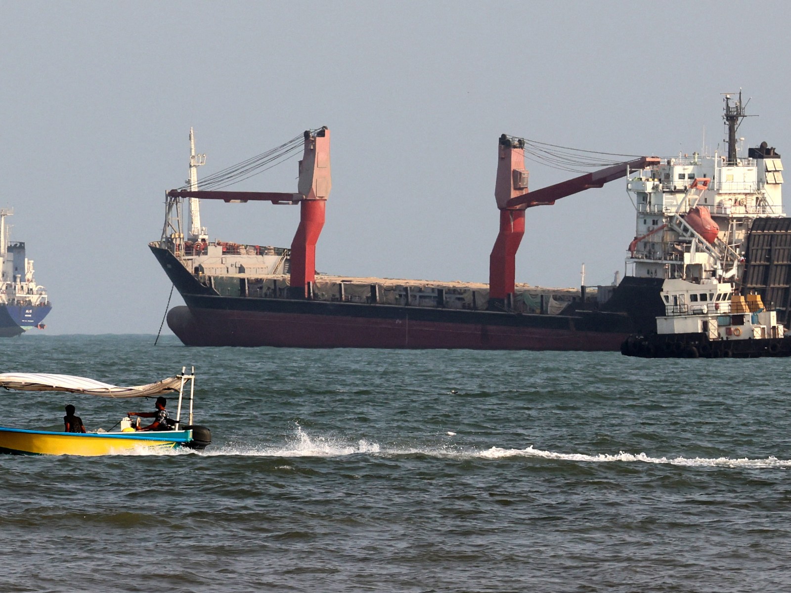 Two commercial cargo ships and a tug lie at anchor while a small open boat with two men aboard speeds past in the foreground