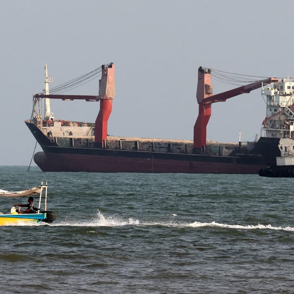 Two commercial cargo ships and a tug lie at anchor while a small open boat with two men aboard speeds past in the foreground