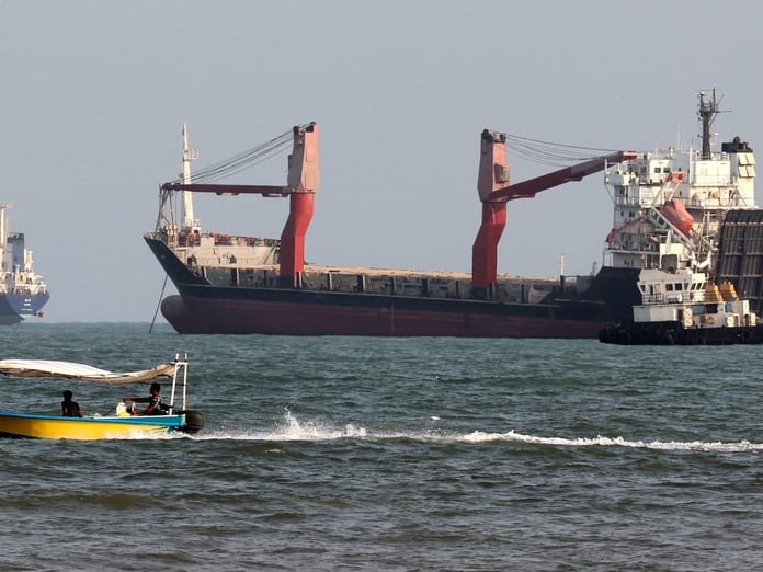 Two commercial cargo ships and a tug lie at anchor while a small open boat with two men aboard speeds past in the foreground