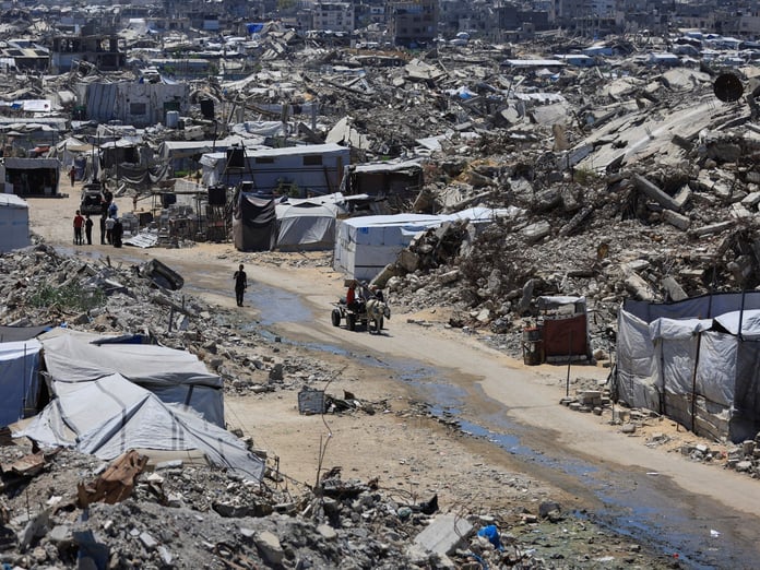 Displacement tents pitched among the rubble of a flattened Gaza neighbourhood, with people and a donkey cart on a dirt track running through it