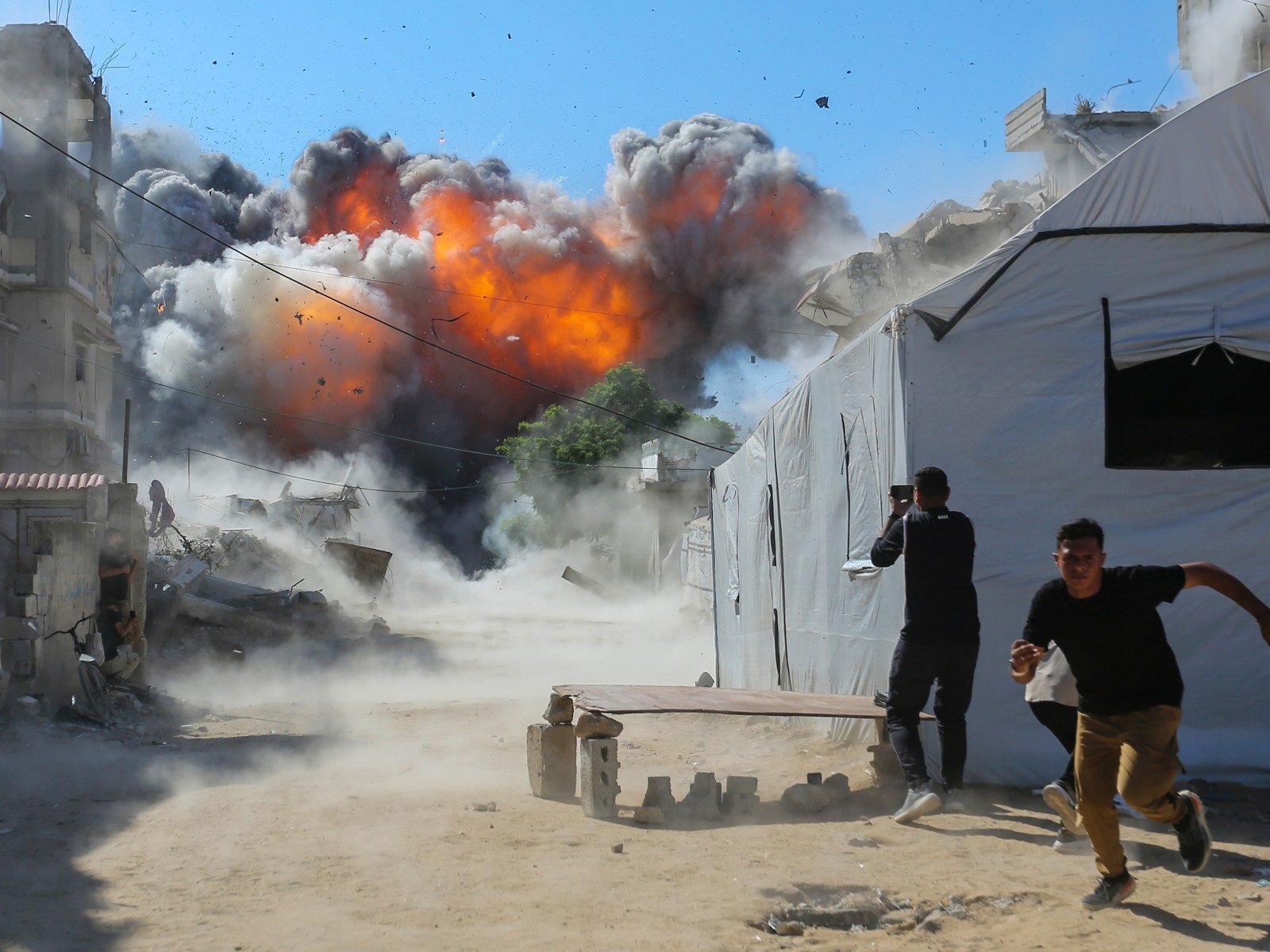 A fireball and smoke rise beside white displacement tents in Gaza as two young men run from the blast