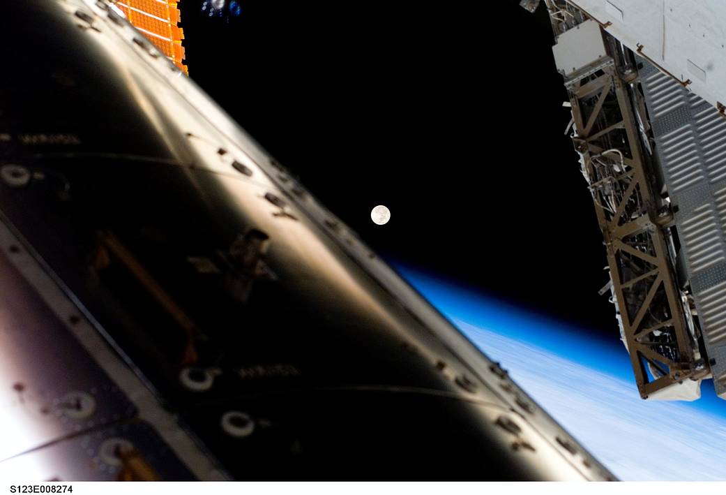 A full moon visible above Earth's horizon and airglow, framed by components of the International Space Station