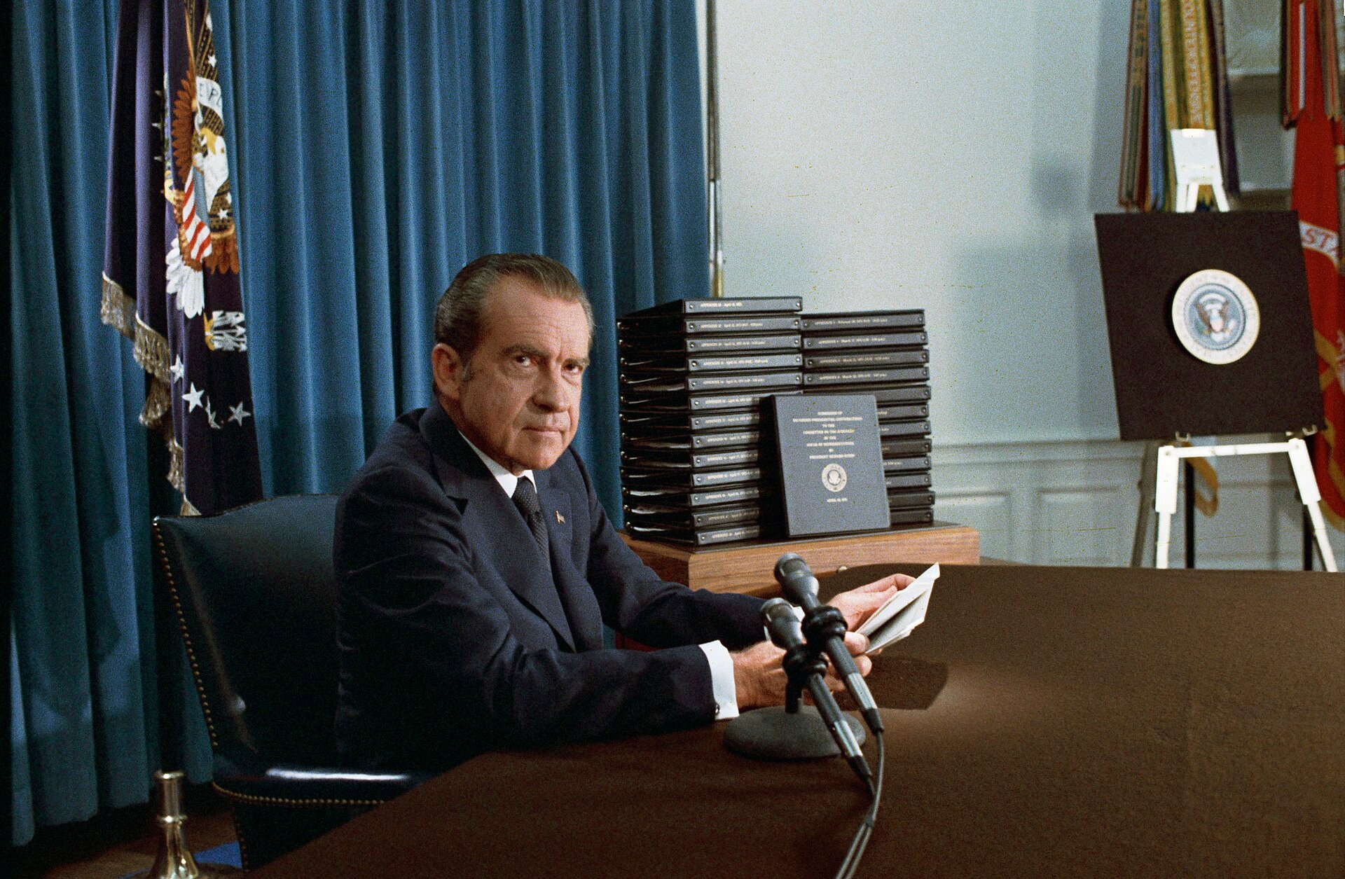 Richard Nixon seated at a desk beside a stack of black binders containing edited transcripts of the White House tapes