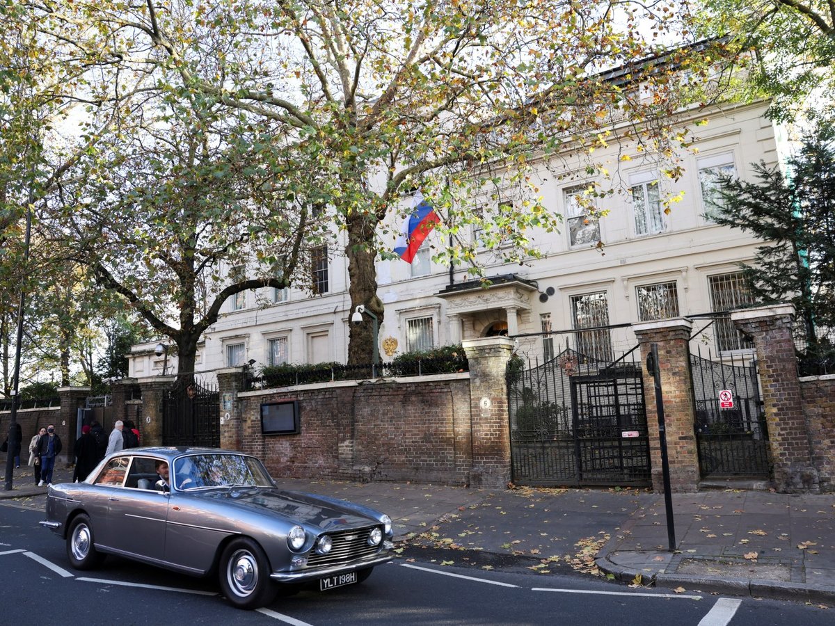 The Russian embassy building at Kensington Palace Gardens in London, with the Russian flag flying above the entrance and a car passing on the road outside
