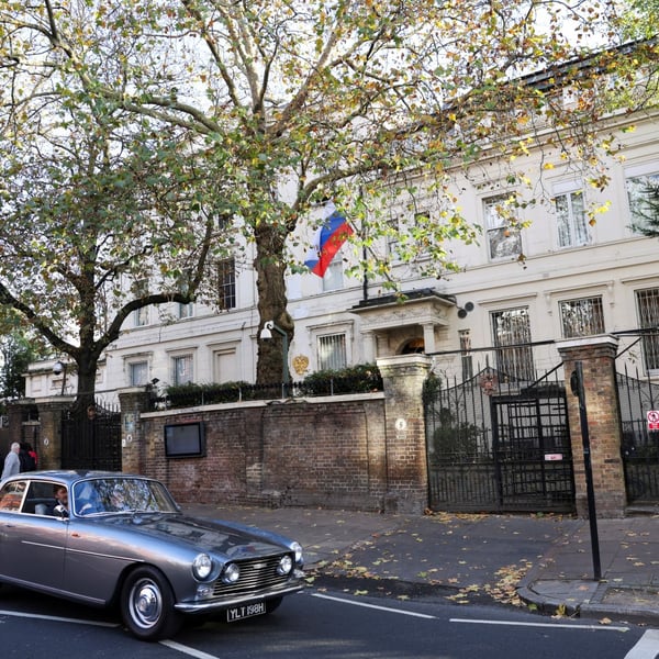 The Russian embassy building at Kensington Palace Gardens in London, with the Russian flag flying above the entrance and a car passing on the road outside