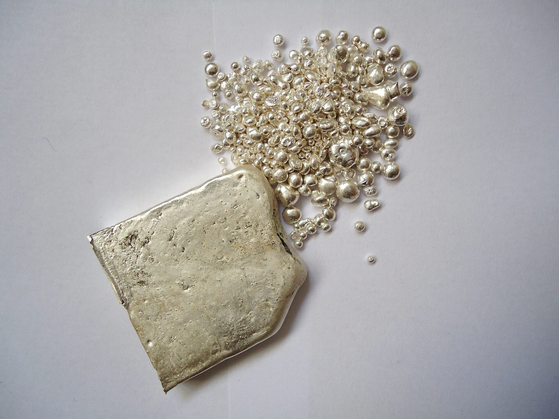 Today silver price: a rough cast silver ingot beside scattered silver casting granules on a white surface