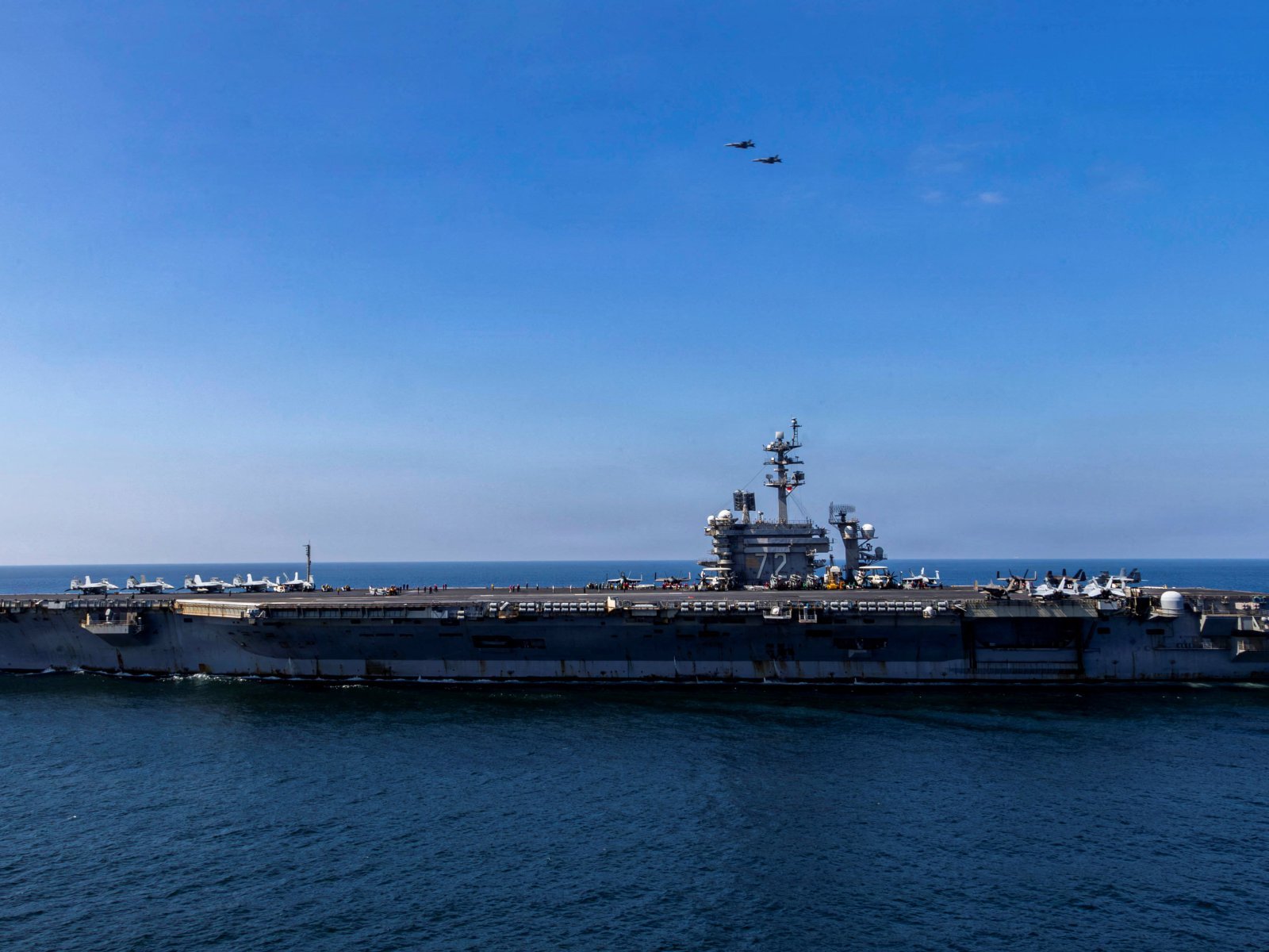 A US Navy aircraft carrier under way with fighter jets ranged along its flight deck and two aircraft flying overhead
