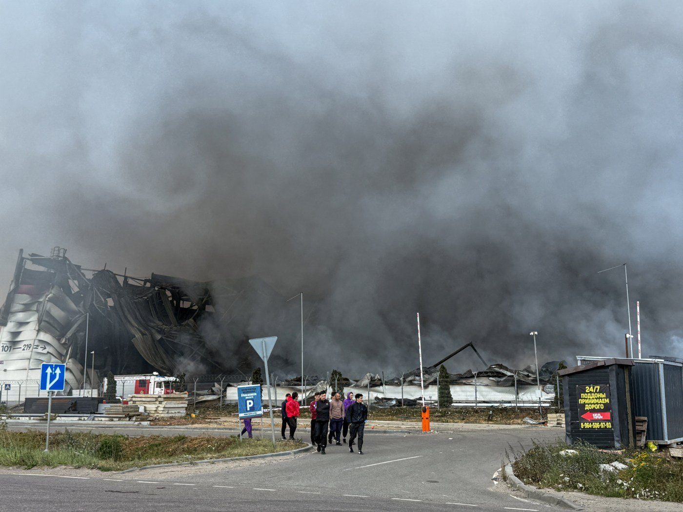 A collapsed warehouse burns behind a security fence outside Moscow, thick black smoke drifting across the road as people walk away from the site