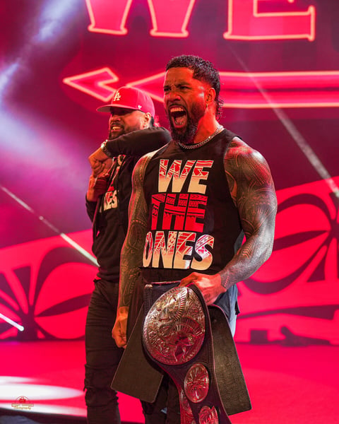 The Usos on the WWE entrance stage in We The Ones shirts, one twin shouting with a tag team title over his shoulder, the other behind him in a red cap