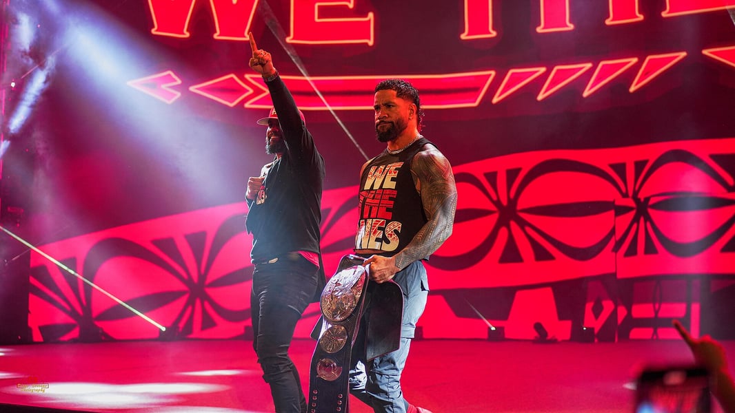 Wide shot of The Usos on the WWE entrance ramp in front of a large red We The Ones screen with Samoan patterning, one twin pointing upward and the other carrying a tag team title