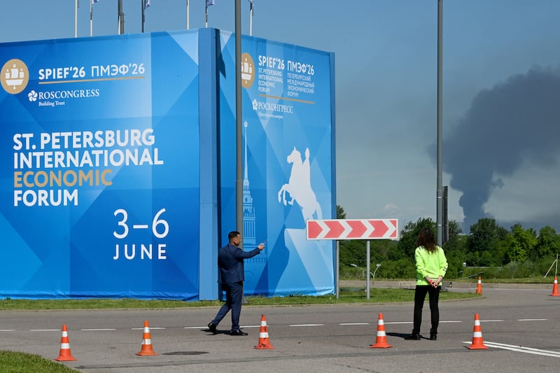 People stand in front of SPIEF 2026 banners as black smoke rises in the distance over St. Petersburg