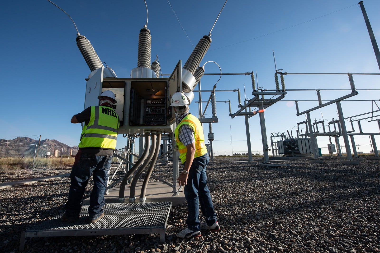 Technicians work at a high-voltage electrical substation with transformers and insulators