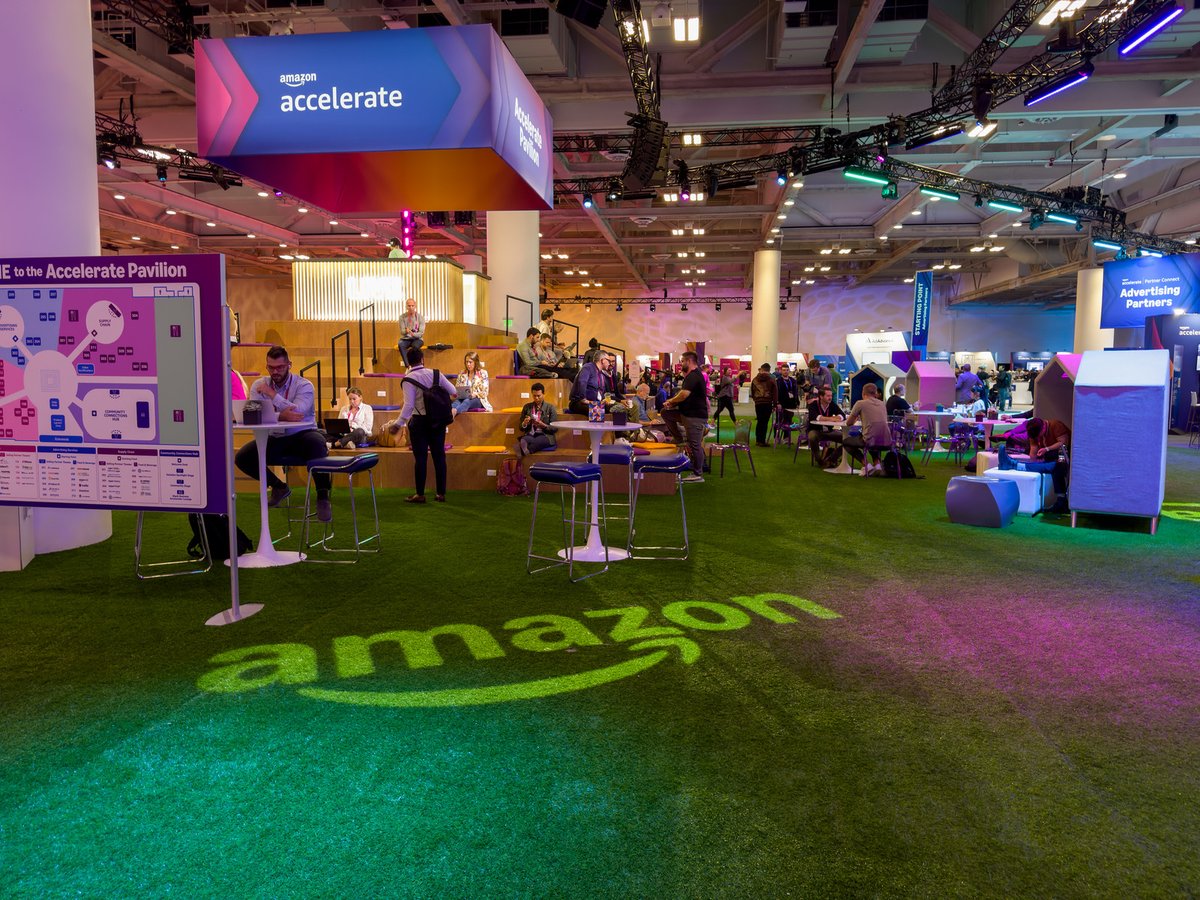 Amazon Accelerate conference pavilion floor showing Amazon logo in green lighting with attendees networking