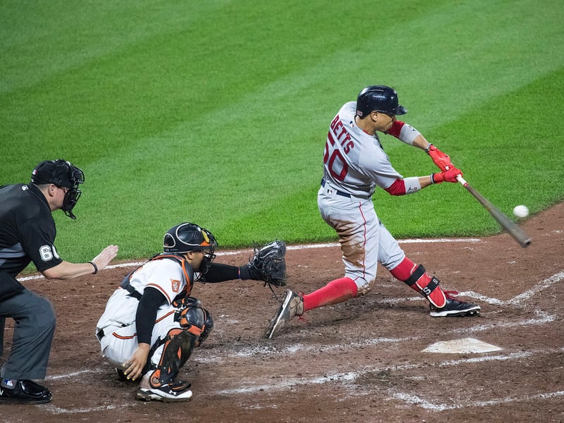 Baseball batter Mookie Betts making contact with the pitch as the catcher reaches for it and the umpire watches from behind