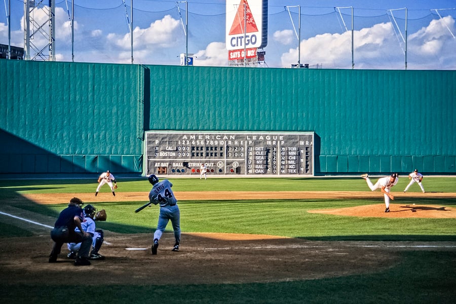 A baseball game at Fenway Park with the pitcher mid delivery, the batter swinging and the manual American League scoreboard on the Green Monster behind them