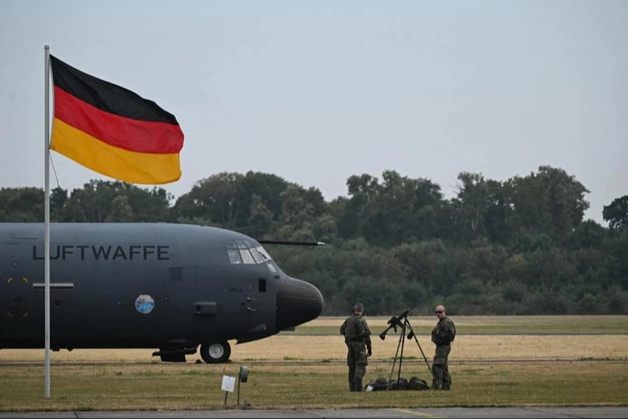 Two Bundeswehr soldiers stand beside a Luftwaffe C-130 transport aircraft on an airfield with the German flag flying