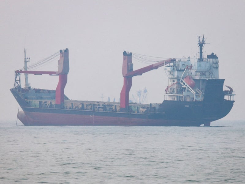 A general cargo ship with red deck cranes lies at anchor in flat grey water and haze near the Strait of Hormuz