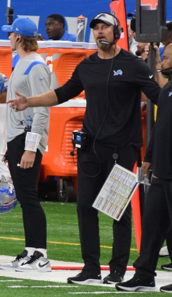 Dan Campbell in black Detroit Lions coaching kit with a headset and play sheet, gesturing on the sideline