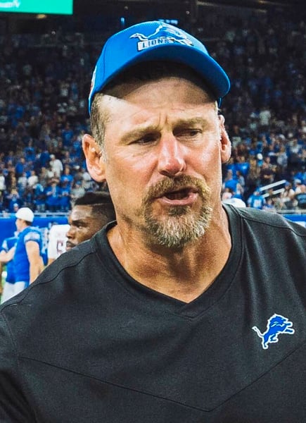 Close-up of Dan Campbell wearing a blue Detroit Lions cap and black Lions shirt on the field