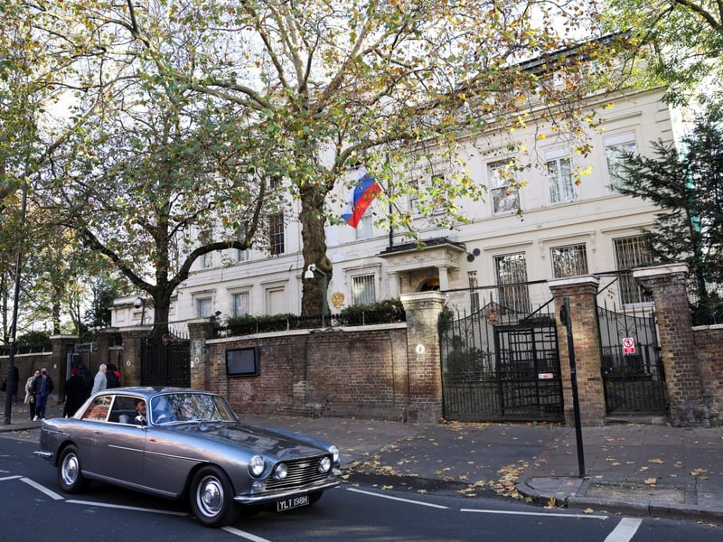 The Russian embassy building at Kensington Palace Gardens in London, with the Russian flag flying above the entrance and a car passing on the road outside