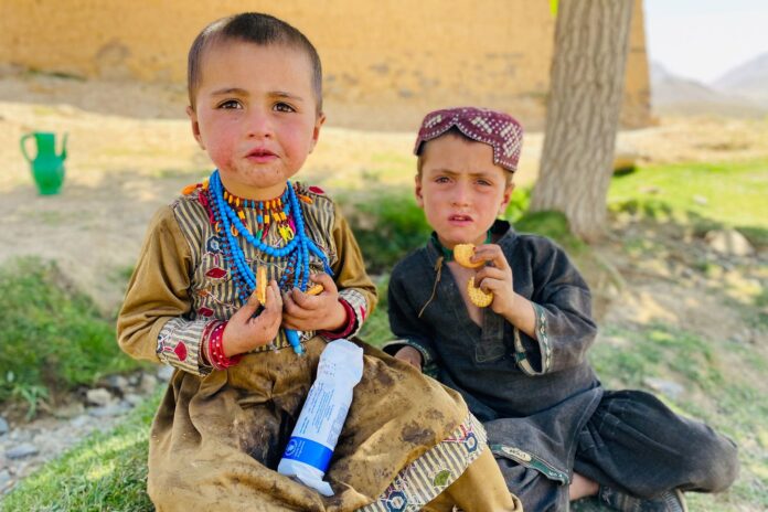 Two young children sit outdoors in Afghanistan eating biscuits, with a WFP-branded package on a girl's lap