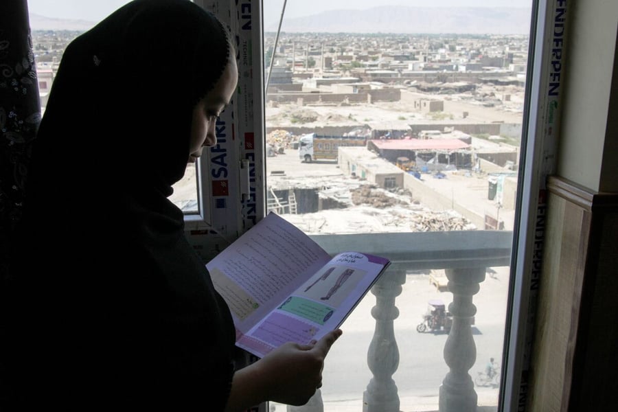 A girl in a black headscarf reads an open book beside a window overlooking a town in Afghanistan