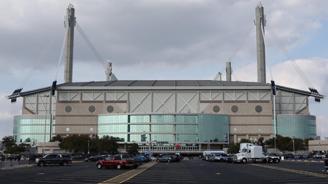 Exterior of the Alamodome in San Antonio under a cloudy sky, with parked cars and trucks in the lot