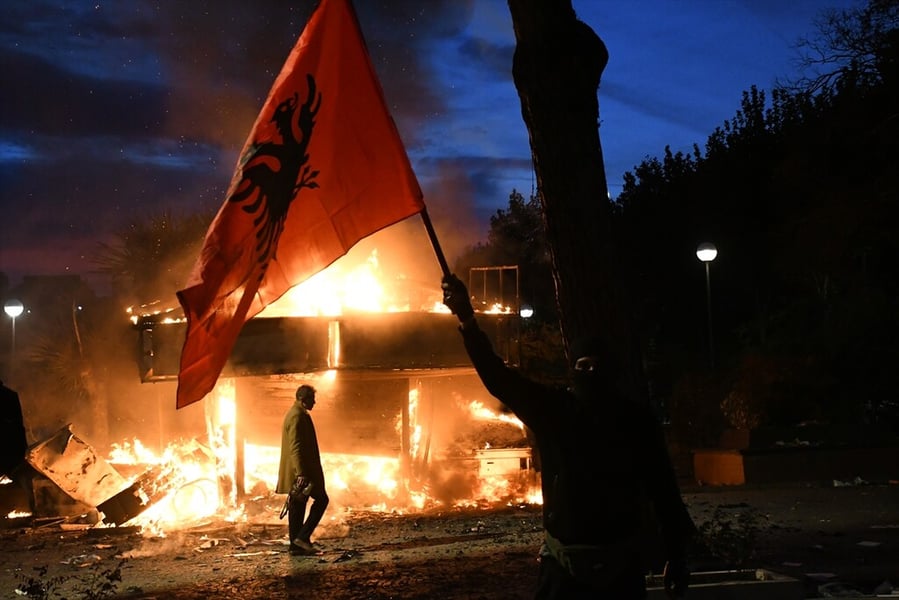 A masked protester raises an Albanian flag in front of a burning structure at night during student protests in Albania