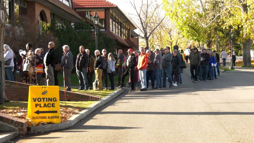 Voters line up along a street beside a yellow Elections Alberta voting place sign during advance voting