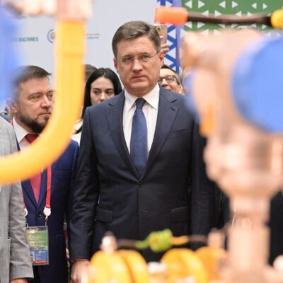 Russia's Deputy Prime Minister Alexander Novak at the Russian Energy Week 2026 forum, standing among attendees with blurred pipe fittings in the foreground