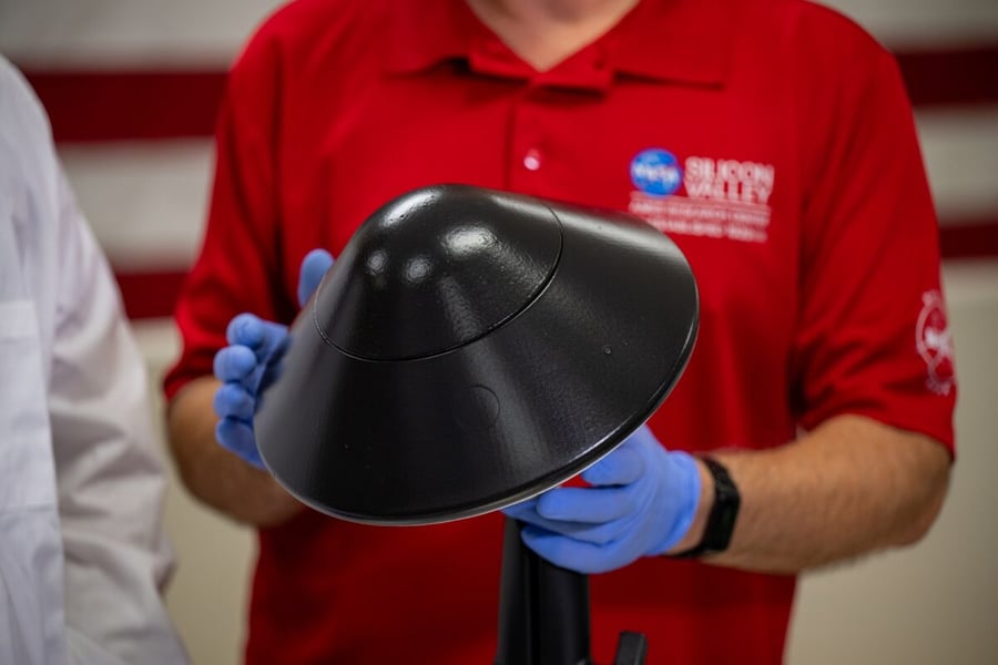 A technician in a red NASA Silicon Valley shirt and blue gloves holds a black cone-shaped heat shield for a KREPE capsule