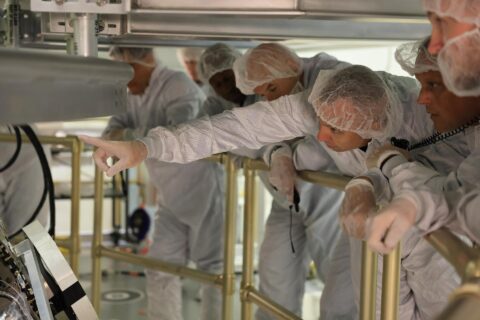 Artemis III astronauts in white cleanroom suits lean over a railing to view the Orion spacecraft at Kennedy Space Center