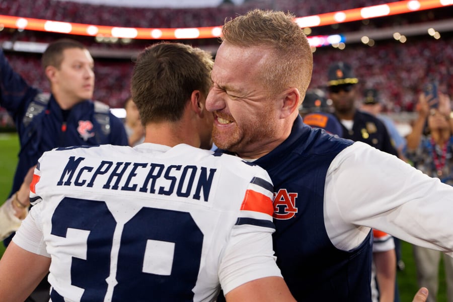 Auburn kicker Alex McPherson in a white jersey embraces a coach in a navy vest on the field in Athens