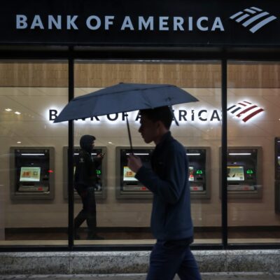 A pedestrian holding an umbrella walks past the illuminated window of a Bank of America branch in New York with a row of ATMs inside