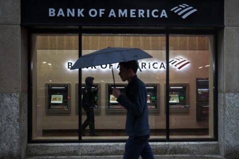 A pedestrian holding an umbrella walks past the illuminated window of a Bank of America branch in New York with a row of ATMs inside