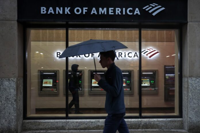 A pedestrian holding an umbrella walks past the illuminated window of a Bank of America branch in New York with a row of ATMs inside