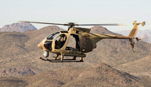 A tan Boeing AH-6i light attack helicopter in flight over desert hills