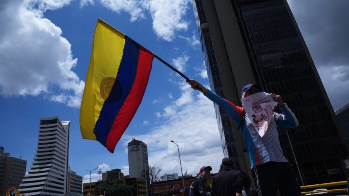 A protester in a blue cap holds up a Colombian flag and a placard against a Bogota skyline